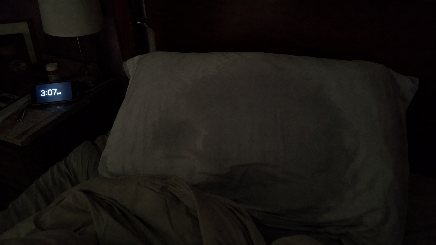 A dim bedroom at night: a rumpled pillow and bedding, a nightstand with a phone glowing 3:07am.